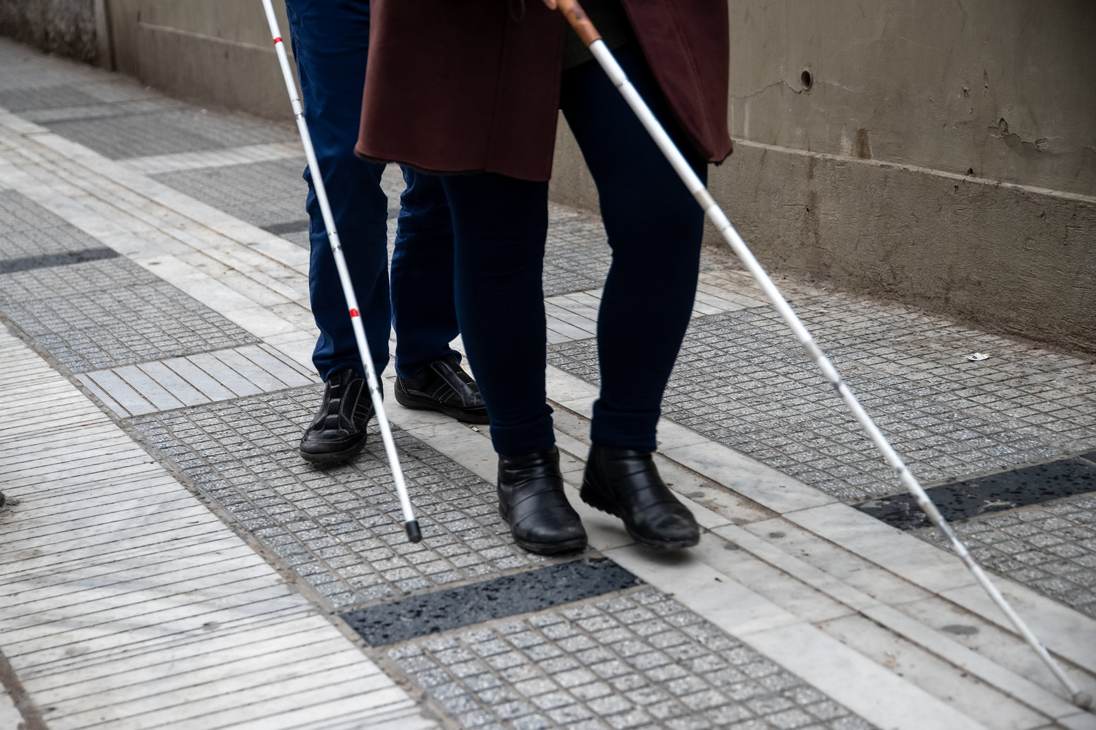 A couple walking down the sidewalk. The view is looking down at their feet and white canes infront of them.